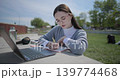A hand-held shot of a young female student in casual clothes and glasses using a laptop and taking notes with a pen in a notebook in an urban park. 139774468