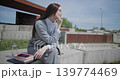 A handheld shot of a young female student in casual clothes sitting on a concrete bench in an urban park, enjoying a sunny summer day. 139774469