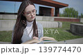 A hand-held shot of a young female student in casual clothes wearing glasses sitting on a concrete bench in an urban park on a sunny summer day and reading a book, flipping through the pages. 139774470
