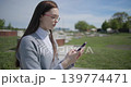 A hand-held shot of a young female student in casual clothes wearing glasses sitting on a concrete bench in an urban park on a sunny summer day using a mobile phone. 139774471