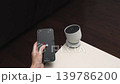 Hand holding smartphone displays app interface while interacting with smart speaker on a table in a modern living room setting 139786200