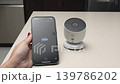Hand holding smartphone connects to a smart home camera on a countertop, displaying connection status and camera view in a modern kitchen setting 139786202