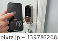 Hand holding smartphone displays locking interface for smart door lock, showcasing the transition from unlocked to locking state in a home setting 139786208