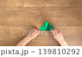 A child's hands folding green origami paper at a wood-grain desk; silent, 4K slow motion. 139810292