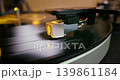 Detailed close-up of a turntable tonearm with white cartridge headshell, positioned over a black vinyl record on the platter, highlighting audio equipment precision. 139861184