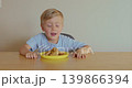 A blond boy with blue eyes is sitting at the table in the kitchen and eating nuggets from a yellow plate 139866394