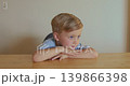 A blond boy is resting his head on his hands on the table, wearing a blue T-shirt, and thinking 139866398