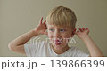 A boy is making funny faces at the camera, holding both hands to his ears and sticking out his tongue. 139866399