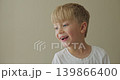 A blonde boy in a white T-shirt is very funny and expressive, sticking out his tongue and looking to the side. 139866400