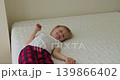 A blonde boy is looking at the camera and smiling while lying on the bed and playing with a small brown toy dog. 139866402