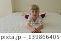 A blond boy in red pajama pants and a white T-shirt is lying on the bed, smiling at the camera, and trying to show different emotions. 139866405