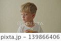 oy with blond hair in a white T-shirt thoughtfully drinks orange juice and looks out the window. 139866406