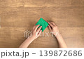 A child's hands folding green origami paper on a wooden desk. 139872686