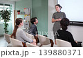 Positive young girl coach standing near interactive board telling new topic or showing presentation with remote control to people sitting on chairs in classroom 139880371