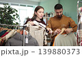 Couple is standing in a European clothing store and holding hangers with dress and basic cotton t shirts. Buying casual clothes at the shopping center 139880373