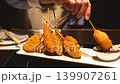 A close-up of a young woman's hands as she generously applies sauce to kushikatsu (deep-fried skewers) at an izakaya (Japanese pub). 139907261