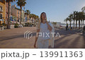 A person strolls along the Villajoyosa coast. The sunlight shines brightly, and palm trees provide shade. The sea is visible in the background as the day begins. 139911363