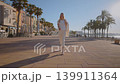 A person walks along the promenade of Villajoyosa, Spain. Palm trees line the path as cafes and shops are seen in the background. The sun shines brightly on this clear day. 139911364