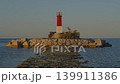 The lighthouse stands on the rocky jetty at Villajoyosa coast. The sun sets in the background, casting warm light over the water and the landscape. 139911386