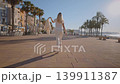 A woman walks along the waterfront in Villajoyosa. The sun is shining bright as she moves past palm trees. The street is quiet, with buildings lining the coast. 139911387