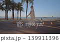 A person walks along the promenade at Villajoyosa coast. The beach and ocean are visible in the background. Palm trees frame the area as the sun begins to set. 139911396