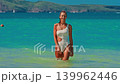 A beautiful girl with a beautiful figure in a white swimsuit sits relaxed on the beach sunbathing and admiring the ocean 139962446