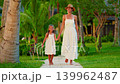 A mother and daughter on vacation, wearing white dresses, stroll hand in hand at sunset along the ocean coast of a tropical island. A beautiful family vacation concept 139962487