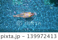 Top down drone view of woman floating in blue swimming pool during sunny tropical holiday and luxury resort relaxation. 139972413