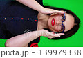 Stylish brunette wearing glasses posing on a green background touching her face with hand. Fashion and optics concept with copy space. 139979338