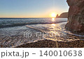 Sunset ocean waves golden sun setting over the sea horizon with foam washing onto a pebble beach by a cliff 140010638