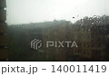 Rain falls on window glass covered with a mosquito net. Peering through, the view shows buildings outside under the gray sky. Water runs down the glass in various patterns 140011419