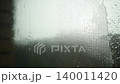 Rain drops hit the glass of a window covered with a mosquito net. Water flows down the surface while clouds cover the sky. The setting shows a typical rainy day at home 140011420