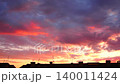 Bright colors fill the sky as the sun sets over a city skyline. The view showcases cloud formations and changing lights during evening time. Rooftops and antennas are visible in the frame 140011424
