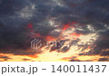 Dramatic clouds fill the sky as the sun sets over the horizon. The colors change from bright orange to deep red and purple. People gather to enjoy the view during summer 140011437