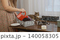Young woman in a floral dress is watering freshly planted seeds in a container with a red watering can, while her cute tabby cat curiously watches the gardening process from the table. 140058305