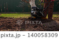 Male farmer using a walk-behind tractor to plow and cultivate the soil in a garden, preparing the land for planting crops during the spring agricultural season with modern machinery. 140058309