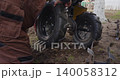 Male farmer in work overalls mounting a new wheel onto the axle of a small walk-behind tractor, performing maintenance on agricultural machinery before working in the muddy field. 140058312