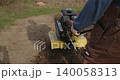 Hardworking farmer in overalls preparing the land for planting by plowing the fertile soil with a powerful motor cultivator, turning up fresh earth in a garden for the new season. 140058313