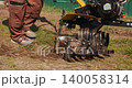 Male worker preparing the soil for planting by plowing the land with a walk-behind tractor in a garden on a sunny spring day, loosening the earth before sowing seeds for cultivation. 140058314