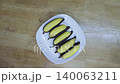 Top down view of five steamed yellow bananas with dark peels on a white square plate. Healthy organic snack on a light wooden table surface. Traditional Southeast Asian style food presentation. 140063211