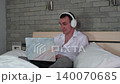 A man is sitting on the bed, using a laptop and wearing headphones. Man listening to music on headphones while working in his bedroom 140070685
