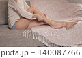 Young woman sitting on a sofa applying moisturizing body lotion on her beautiful legs. Skincare routine and cosmetic body care concept 140087766