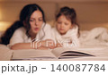 Loving mother reading a fairytale book to her cute daughter before bedtime. Happy family spending quality time together in a cozy bedroom, focus on book 140087784