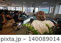 Group of diverse individuals seated in a modern office space attentively watching a presentation with a speaker visible in the background, surrounded by greenery and soft seating 140089014