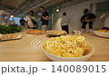 Group of people socializing in a modern office setting with a bowl of potato chips in the foreground and various snacks on a wooden table in the background 140089015