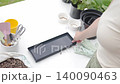 Woman prepares flower pot for container gardening, placing a tray and black planter on a covered table. Concept of planting setup and DIY plant care 140090463