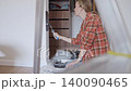 Woman applies black paint to white wall with small roller beside a tray, bucket, and protective sheet. Renovation and DIY decorating concept 140090465