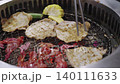 Traditional Japanese yakiniku barbecue with slices of beef, pork, and corn grilling on a charcoal mesh. Metal tongs and chopsticks are used to flip the meat over glowing red embers. 140111633