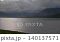 Drone flies back over grass shore as light shafts illuminate shore of Loch Linnhe on a spring evening with stormy clouds in sky 140137571