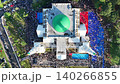 An aerial view of the Baitul Faizin Grand Mosque during the busy Eid al-Fitr prayer, with the signature green dome and twin minarets, a vast congregation on blue mats 140266855